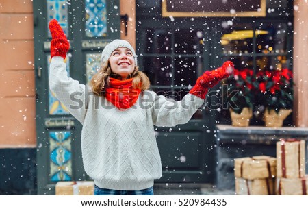 Young pretty woman near the shop with gifts enjoying winter and  looking up on falling snow.
Girl dressed  in warm white hat, red scarf and mittens.
Happy holiday  and freedom concept.