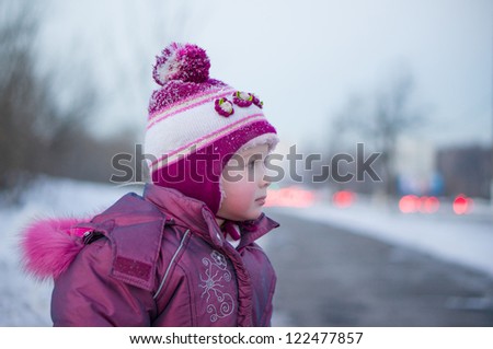 Adorable girl walking and playing in winter park in evening. Look at road with moving cars on back