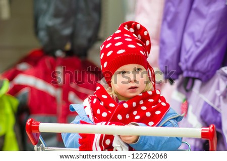 Adorable baby on cart choose clothes in supermarket. Try on red winter hat and scarf