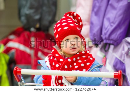 Adorable baby on cart choose clothes in supermarket. Try on red winter hat and scarf