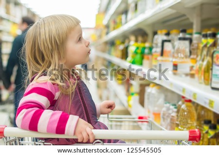 Adorable girl in shopping cart selecting sunflower oil in supermarket