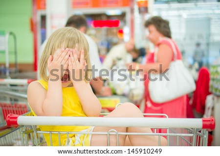Adorable girl sit in shopping cart and close face with hands near cash desk