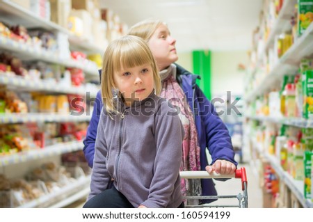 Mother with daughter in shopping cart select products on shelves in supermarket