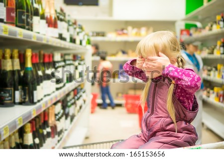 Adorable girl grimacing sitting in shopping cart in alcoholic beverages department in supermarket
