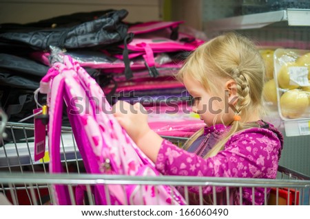 Adorable girl at shopping cart select pink school bag in supermarket