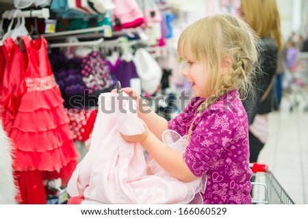 Adorable girl on shopping cart select pink dress in supermarket