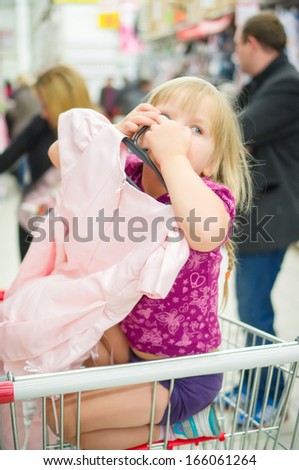 Adorable girl on shopping cart select pink dress in supermarket