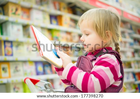 Adorable girl sit on shopping cart with kids book in supermarket