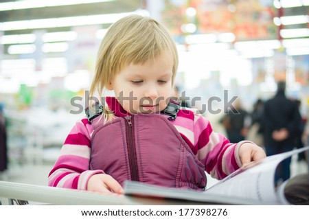 Adorable girl sit on shopping cart and read magazine in supermarket