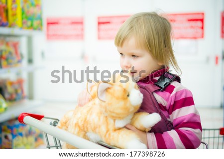Adorable girl sit on shopping cart with plush kitten toys in supermarket
