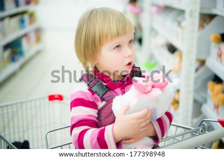 Adorable girl sit on shopping cart with plush kitten toy in supermarket