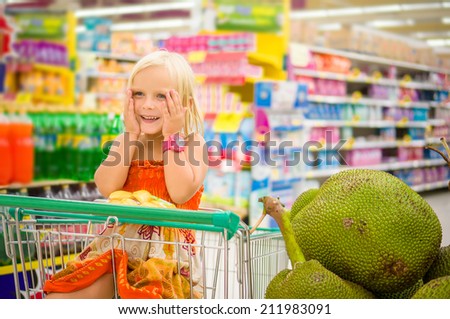 Adorable girl in shopping cart looks at giant jack fruits on boxes in supermarket