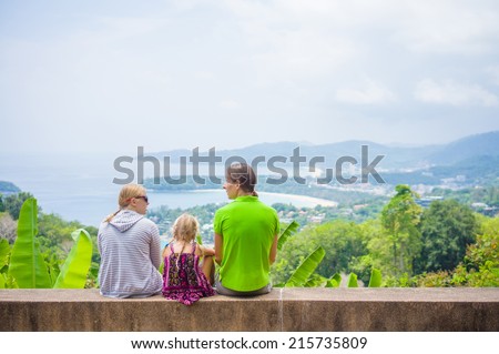 Mother, father and daughter sit on side and look around from mountain view point on tropical beach island
