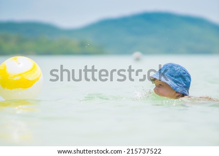 Adorable girl in blue hat swim in ocean near beach. Play with yellow ball