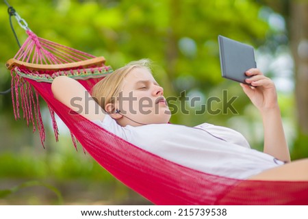 Nice young lady listen music and read book on e-ink reader in  hummock under palm trees on tropical beach. Close up