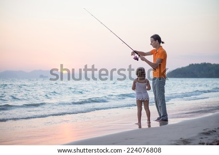 Father with adorable daughter fishing with rod on ocean beach on sunset