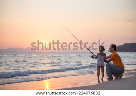 Father with adorable daughter fishing with rod on ocean beach on sunset