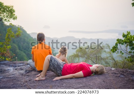 Father and daughter  seat, mother lie near cliff on the top of tropical island mountain on sunset