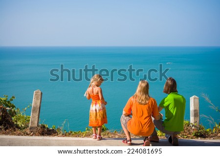 Young mother, father crouch and daughter stay on mountain road along tropical island beach and look at ocean