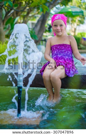 Adorable girl in violet dress sit on bridge above water stream and fountain in front