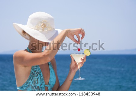 Photo of a beautiful young woman in a hat by the sea holding a fresh cocktail