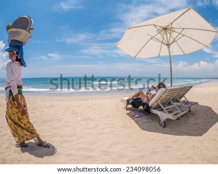 Lady in white hat sitting in chaise longue in Bali with old beach seller passing by
