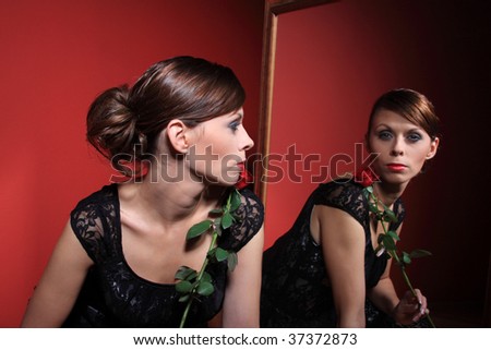 A beautiful sexy women wearing an evening dress and holding a red rose looking at her reflection in the mirror on red background