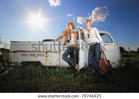 A young couple on a old broken car in a field man is holding a leather bag