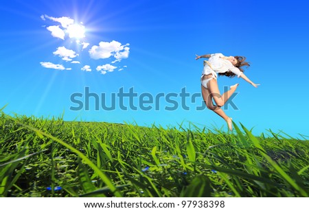 Young woman jumping on a green grass hills under midday sun in blue sky.