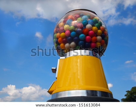 A vintage yellow gumball machine with multi-colored gumballs stands tall in front of a blue sky with wispy clouds.