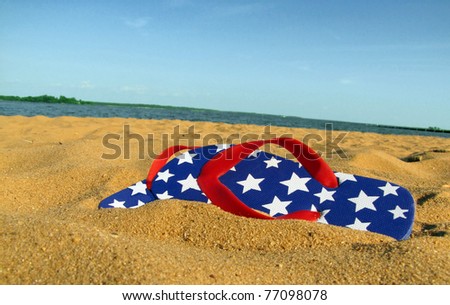 A pair of red, white, and blue flip flops with stars, on a sandy beach with water and sky in the background.