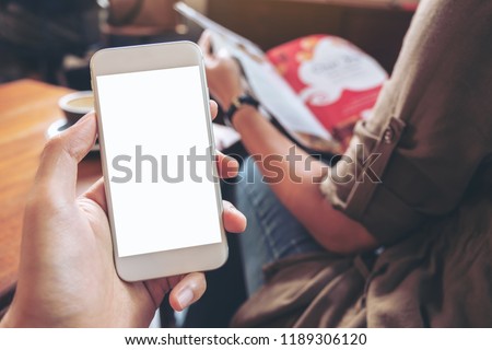 Mockup image of hand holding white mobile phone with blank screen with woman reading books in modern cafe