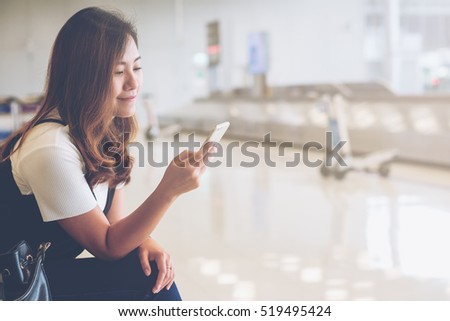 A beautiful asian woman using mobile phone with feeling happy and smiley face , sitting and waiting for baggage claim in blur airport background