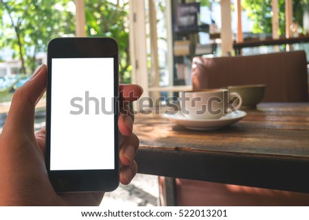 Mockup image of hand holding black mobile phone with blank white screen and hot latte coffee on vintage wood table in cafe