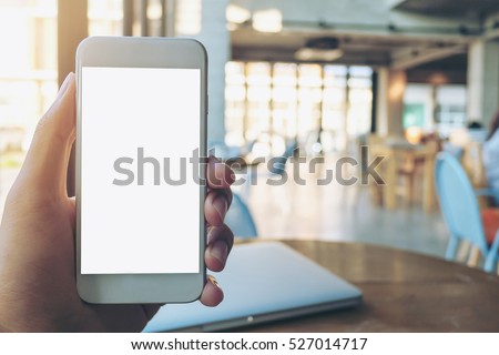 Hand holding white mobile phone with blank white screen and silver laptop on vintage wood table in cafe