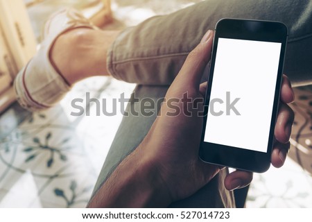 Mockup image of hand holding black mobile phone with blank white screen on thigh with white canvas shoes\
 at vintage tile floor in cafe , feeling relax