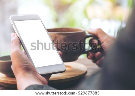 Mockup image of left hand holding white mobile phone with blank white screen and Right hand holding hot latte art coffee cup while looking and using it at vintage wooden table in cafe