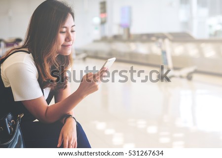 A beautiful asian woman using mobile phone with feeling happy and smiley face , sitting and waiting for baggage claim in blur airport background