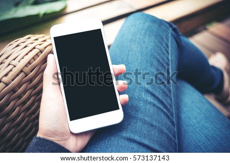 Mockup image of hand holding white mobile phone with blank black screen on thigh