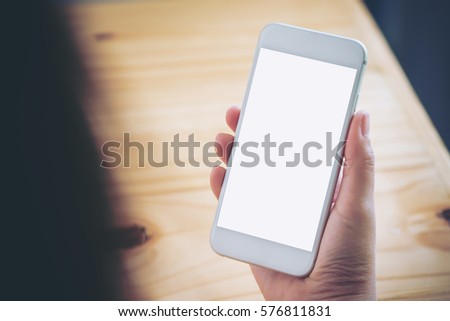 Mockup image of hand holding white mobile phone with blank white screen on vintage wood table in cafe