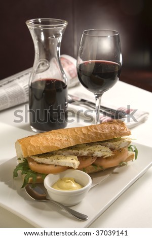 French loaf cheese sandwich with red wine in a glass and caraffe, with camembert cheese, tomato and lettuce on the sandwich, with Dijon mustard. A newspaper is out of focus in the background