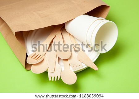 Eco-friendly disposable utensils made of bamboo wood and paper on a green background. Draped spoons, fork, knives, bamboo bowls with paper cups and packet