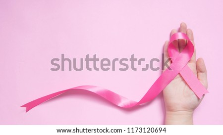 International symbol of Breast Cancer Awareness Month in October. Close up of female hand holding satin pink ribbon awareness on pink background w/ copy space. Women\'s health care and medical concept.
