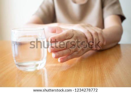 Elderly woman hands w/ tremor symptom reaching out for a glass of water on wood table. Cause of hands shaking include Parkinson\'s disease, stroke or brain injury. Mental health neurological disorder.