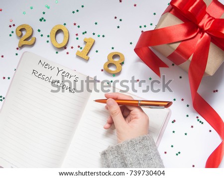 2018 New Year\'s Resolutions concept. Top view of women hands writing goal for new year or Christmas on opened blank notebook with gift box and party decorations on white table. Copy space. Flat lay.