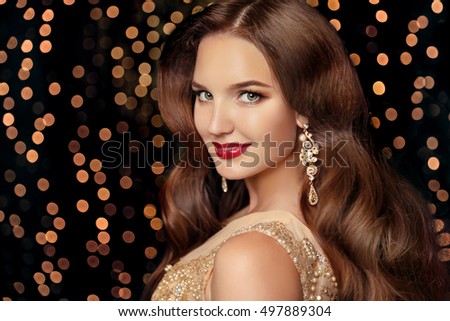 Makeup. Elegant hairstyle. Beautiful brunette smiling with long wavy hair, red lips makeup and fashion jewelry earrings. Attractive girl isolated on holiday party lights background.