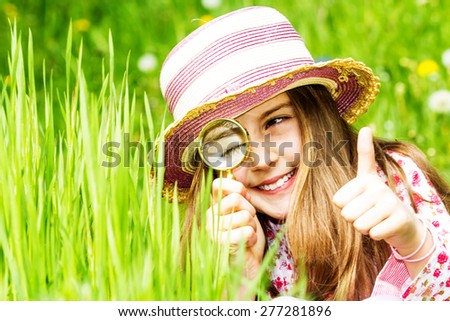 Little girl is playing on green meadow and examining field flowers using magnifying glass