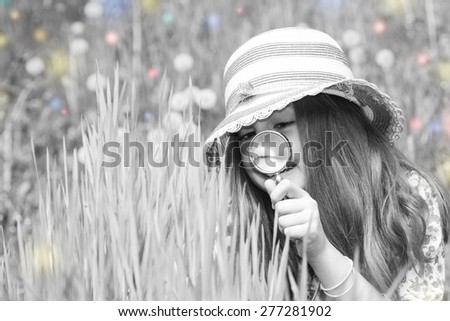 Little girl is playing on green meadow and examining field flowers using magnifying glass