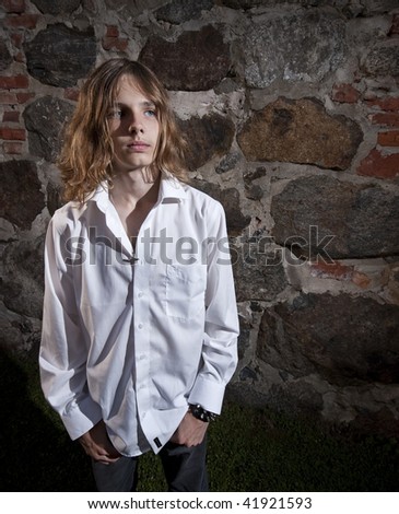 A teenage male model with long hair posing in front of a stone wall.