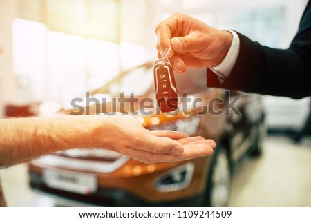 Keys from new car, young salesman transfer the car key in hand of customer in casual wear on dealership background